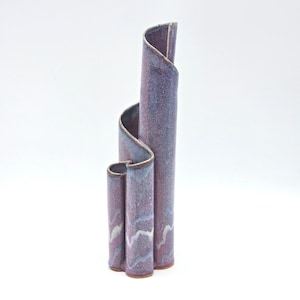 May include: A tall, abstract ceramic vase in shades of purple, blue, and white. The vase has a unique, curved design with three vertical sections that twist and turn. The ceramic has a speckled texture and a glossy finish.