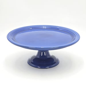 May include: A cobalt blue ceramic cake stand with a wide, round top and a pedestal base. The stand has a glossy finish and is suitable for displaying cakes, cupcakes, or other baked goods. The stand measures approximately 12 inches in diameter.