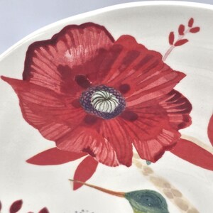 May include: A white ceramic plate featuring a hand-painted design of a red poppy flower. The poppy has a dark purple centre and is surrounded by red petals. A hummingbird is also visible.