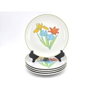 May include: Set of four white ceramic plates with a floral design. Each plate features a colorful illustration of yellow, blue, and orange flowers with green stems. The plates have a green border and are displayed on black plate stands.