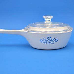 May include: White saucepan with a clear glass lid and handle. The pan features a blue floral design on the side. The handle extends to the left. The background is a solid blue.