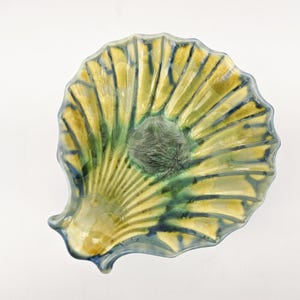 May include: A ceramic seashell-shaped dish with a textured surface. The dish features a yellow and green color scheme with blue accents. The center of the dish has a darker, textured area.
