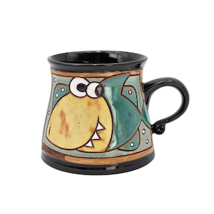 May include: A ceramic mug with a whimsical shark design. The mug features a black handle and rim, with a yellow shark body and a teal fin. The design is outlined in black and brown, with white accents.