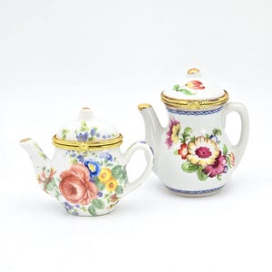 May include: Two small, white porcelain teapots with floral designs. One teapot features a large red rose, while the other has a mix of colorful flowers. Both have gold-colored accents and lids.