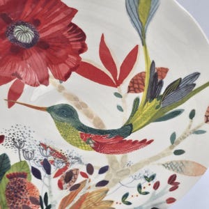 May include: A white ceramic plate with a watercolour design featuring a red poppy, a hummingbird, and various floral elements in shades of red, green, yellow, and blue. The plate has a slightly irregular shape.