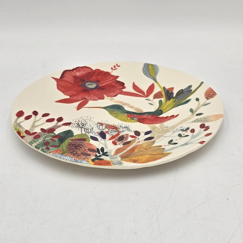 May include: A cream-coloured ceramic plate with a floral and hummingbird design. The plate features a large red flower, a hummingbird, and various other flowers and leaves in shades of red, green, and orange.