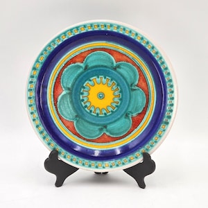 May include: A decorative ceramic plate featuring a central turquoise flower design with a yellow center, set against a red background. The plate has a dark blue border and a white rim with yellow and turquoise square accents. The plate is displayed on a black stand.