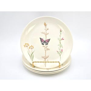 May include: A set of four cream-colored ceramic plates. Each plate features a central design of a pink and black butterfly with floral and botanical accents in yellow and pink. The plates are displayed on a gold-colored wire stand.