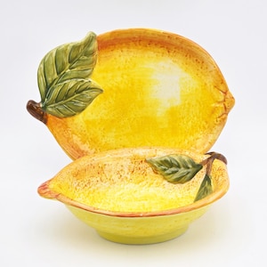 May include: A ceramic lemon-shaped serving dish and lid set. The dish and lid are yellow with brown accents and green leaves. The set is designed to resemble a lemon, with a textured surface.