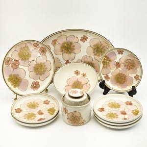 May include: A collection of vintage floral-patterned ceramic dishware. The set includes plates, bowls, and serving platters, all adorned with a repeating pink and yellow flower design. The edges of the dishes have a brown trim. The set is displayed on a white background.