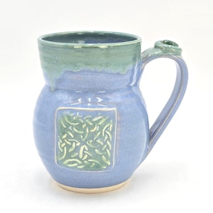 May include: A handmade ceramic mug with a blue body and a green rim and handle detail. The mug features a square, raised design in green with an interwoven pattern. The mug is designed for hot or cold beverages.