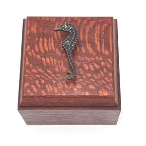 May include: A square wooden box with a dark brown finish and a raised, textured grain pattern on the lid. A metallic, dark bronze-colored seahorse figurine is attached to the center of the lid, serving as a decorative handle or accent piece.