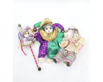 May include: Three decorative clown dolls with porcelain faces and colourful fabric outfits. One doll holds a staff. The clowns wear green, purple, gold, and patterned trousers. The dolls are on a white background.