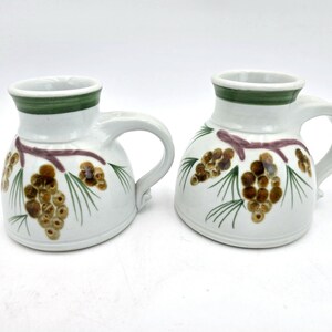 May include: Two white ceramic mugs with green rims and brown pine cone designs. The mugs have handles and are decorated with brown pine cones and green pine needles.