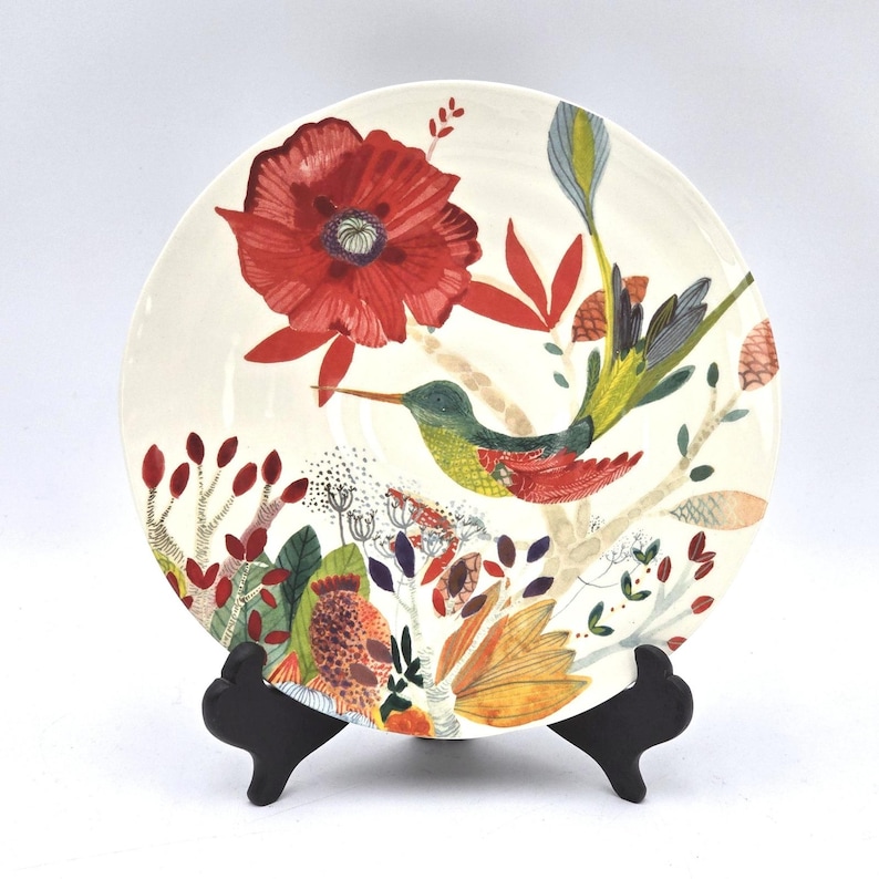 May include: A decorative ceramic plate with a floral and hummingbird design. The plate features a large red poppy, colourful foliage, and a hummingbird with green, yellow, and red feathers. The plate is displayed on a black stand.