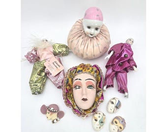 May include: A collection of vintage decorative items, including porcelain dolls, a face mask, and small decorative masks. The dolls are dressed in pink and gold outfits. The face mask has a painted face with detailed features. The items are arranged on a white surface.