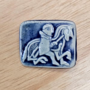 May include: A rectangular ceramic brooch with a glossy finish featuring a dark blue and off-white glaze. The surface displays a relief design of a figure riding a horse, with the horse depicted in profile and the rider facing forward.