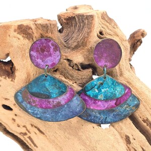 May include: A pair of dangle earrings featuring a circular top piece and a tiered, fan-shaped bottom section. The metal surfaces display a mottled, weathered finish in vibrant shades of magenta, teal, and deep blue, creating a textured appearance.