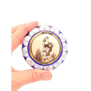 May include: A round metal pill box featuring a central circular enamel portrait of a child carrying a basket. The border is decorated with intricate blue and white floral patterns and gold accents. The lid is hinged and the metal has a polished finish.