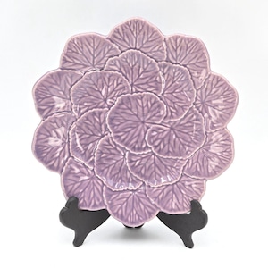 May include: A lavender-colored decorative plate with a leaf design. The plate is shaped like a flower, with overlapping leaves. The plate is displayed on a black stand against a white background. The plate is made of ceramic.
