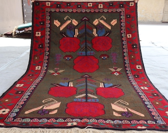 May include: Hand-woven rug with a central olive-green field featuring red and blue floral motifs. The border is red with floral and geometric patterns. The rug has a fringed edge and is displayed outdoors.