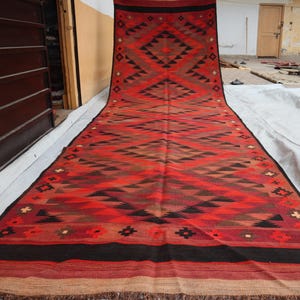 May include: A red, black and brown patterned rug with a geometric design. The rug is made of wool and has a fringed border.