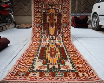 May include: A rectangular, hand-woven rug with a central geometric design in shades of brown, yellow, and red. The rug features a cream-colored background with intricate border patterns and fringed edges. The rug is displayed on a white surface.