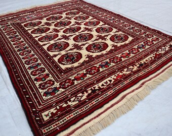 May include: A traditional rug with a cream background and a red, black, and white geometric pattern. The rug has a fringe border.