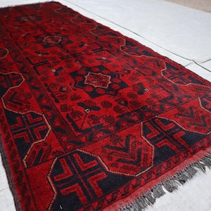 May include: A rectangular, hand-woven rug with a predominantly red field and intricate geometric patterns in black and gold. The rug features a central medallion and border designs, with fringed edges. The rug's dimensions are not visible.
