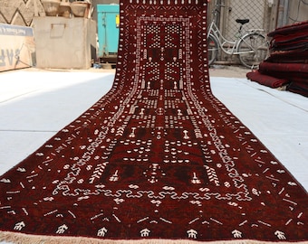Puede incluir: Una alfombra roja larga y estrecha con un patrón geométrico blanco. La alfombra tiene flecos en ambos extremos.