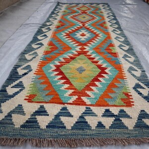 Peut inclure: Un tapis coloré avec un motif géométrique dans des tons de bleu, rouge, orange, vert et blanc. Le tapis a une bordure à franges.