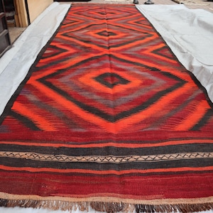 May include: A long, rectangular rug with a geometric pattern in shades of red, orange, black, and gray. The rug has a fringe border and is woven with a traditional design.