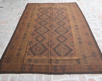 May include: A rectangular woven rug featuring a repeating geometric diamond pattern in shades of dark brown and tan. The rug has a central panel with larger diamonds and a border with smaller motifs, finished with short white fringe at the ends.