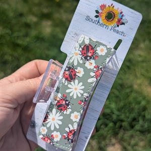 May include: A clear plastic hair claw clip with a green background featuring red ladybugs and white daisies. The clip is attached to a white and gray Southern Peach branded tag with a floral design.