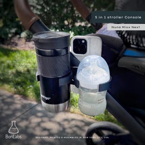 May include: A black 5 in 1 stroller console for Nuna Mixx Next, featuring a black Contigo travel mug, a white smartphone, and a clear Philips Avent baby bottle with measurement markings. The console is attached to a stroller handle.