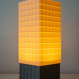 May include: A rectangular table lamp featuring a grid-patterned structure. The upper portion is translucent orange and emits a warm glow, while the lower base section is solid black with a matching grid texture on all visible sides.
