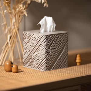 May include: A square tissue box with a textured, geometric design in a neutral colour. White tissues are visible at the top. Decorative wooden figurines and dried plants in a glass vase are also present on a wooden surface.