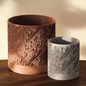 May include: Two cylindrical planters, one brown and one gray, with a geometric pattern. The brown planter has the text "FORAGE" printed on the bottom.