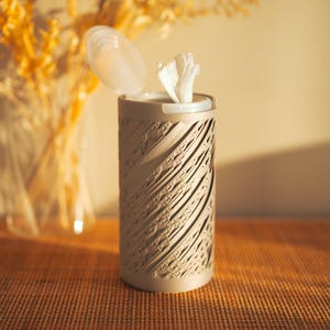 May include: A cylindrical tissue dispenser with a textured, carved design in a neutral color. The lid is open, revealing white tissues. The dispenser sits on a woven surface, with a blurred background of dried flowers.