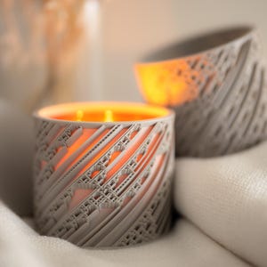 May include: Two lit, gray candles with intricate, geometric designs. The candles sit on a white surface, with the warm glow of the flames visible through the cut-out patterns. The overall aesthetic is modern and elegant.