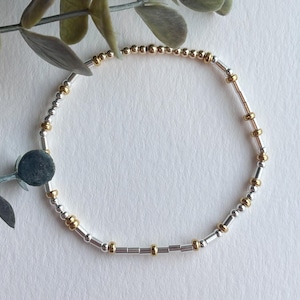 May include: A delicate bracelet featuring a mix of silver and gold beads. The bracelet has a flexible design, with small silver and gold beads interspersed throughout. The bracelet is shown on a white surface with green leaves in the background.