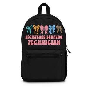 Puede incluir: Mochila negra con un bolsillo frontal con cremallera y un asa superior. La mochila presenta lazos coloridos y el texto "REGISTERED BEHAVIOR TECHNICIAN" en rosa y blanco. Los lazos son en tonos azul, dorado, rosa, azul y estampado de leopardo.