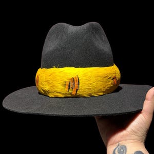May include: A black fedora hat with a wide brim. The hat features a bright yellow band around the crown, adorned with orange and black feather accents. The hat is held against a black background.