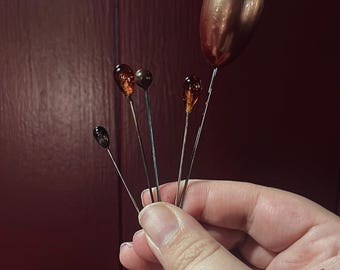 May include: A collection of decorative pins with various jewel-toned, oval-shaped heads. The pins are held in a hand against a dark red background. The heads of the pins range in color from dark brown to amber and copper.