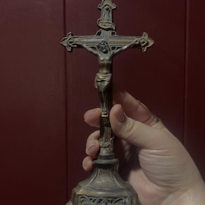 May include: A bronze-colored crucifix with a detailed depiction of Jesus on the cross. The cross is mounted on a decorative base. The religious item is held in a hand against a dark red background.