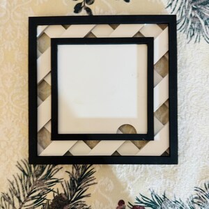 May include: Square black picture frame with a white mount and a woven cream and black border. The frame has a clean, modern design, suitable for displaying photos or artwork. The frame is sitting on a floral patterned surface.