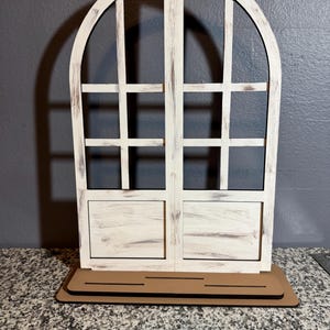 Peut inclure: Un cadre de fenêtre en bois blanc vieilli avec un motif de grille, posé sur une base marron. Le cadre a un aspect rustique et patiné, avec le grain du bois visible et une conception à deux portes. L'esthétique générale est de style ferme ou cottage.