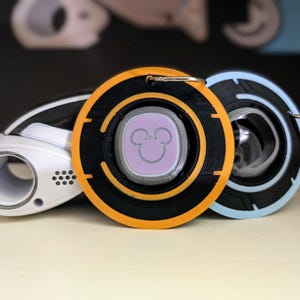 May include: A black and orange round keychain with a Mickey Mouse icon in the center. The icon is a light purple color and is surrounded by a black ring. The keychain is attached to a white and blue round keychain.