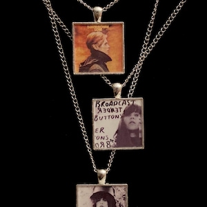 Peut inclure: Trois colliers en argent avec des pendentifs carrés représentant des pochettes d'album. Le pendentif du haut représente la pochette de l'album "Low" de David Bowie, le pendentif du milieu représente "Broadcast" de "The Tender Buttons" et le pendentif du bas représente la pochette de l'album "T. Rex".
