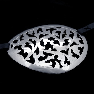 May include: A silver eye patch with a decorative, intricate, floral design. The patch is made of metal and has a black elastic strap.